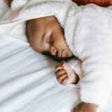 An infant quietly sleeping on a white mattress with an orange embroidered pillow behind him. The baby is in a white fuzzy jumper with little bear ears on top of the hood.